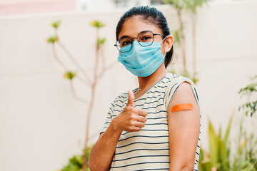 Portrait of an Indian teen giving thumbs up after getting vaccinated and showing bandage on his arm. with wearing protective mask - obrazy, fototapety, plakaty