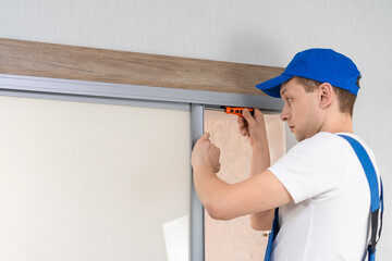 Handyman who installs sliding doors at home in overalls with a screwdriver in his hands repairs the...