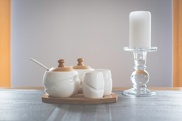 Cozy home concept. White porcelain seasoning containers for salt, pepper, sugar with wooden details. Big candle on transparent stove from glass. Selective focus on small container, blurred background.
