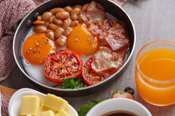 Traditional full English breakfast in cooking pan with fried eggs, bacon, beans, grilled tomatoes. toast, butter, jam on a wooden board, coffee and orange juice on stone background. top view.