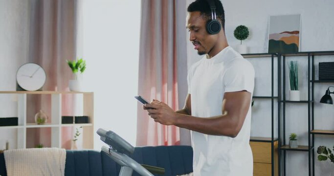 Likable smiling in good form sporty black-skinned guy in headphones walking on running track at home and applying his phone to chat with friends