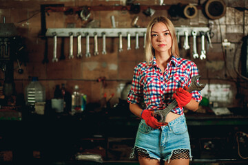 A beautiful young girl in a plaid shirt and short denim shorts with a wrench in her hands at a service station. Model in orange gloves in the garage.