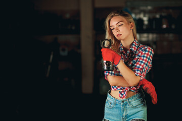 A beautiful young girl in a plaid shirt and short denim shorts holds a piston-connecting rod of the...