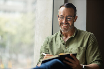 Mature man with beard, glasses writing in book sitting by sunny window - Powered by Adobe