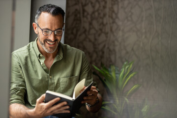 Mature man reading book by window, smiling wearing glasses, copy space - obrazy, fototapety, plakaty