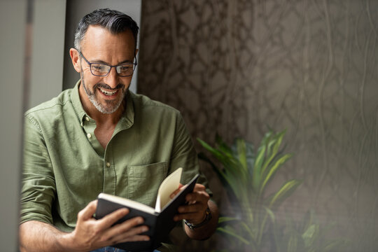Mature man reading book by window, smiling wearing glasses, copy space