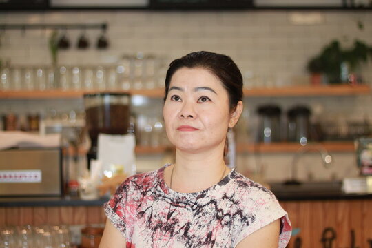 Asian 40s Woman Looking At The Food And Drink Menu On The Board Above.in The Coffee Shop In The Morning.Selective Focus At Face .food And Drink  Concept.