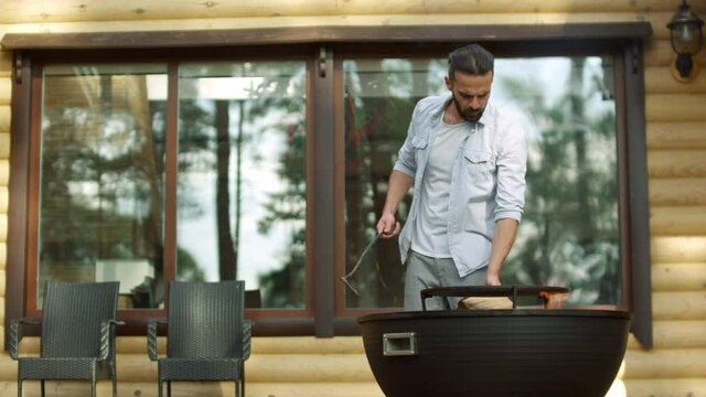 Focused guy using poker outdoors. Man chef cooking food on bbq grill near house.