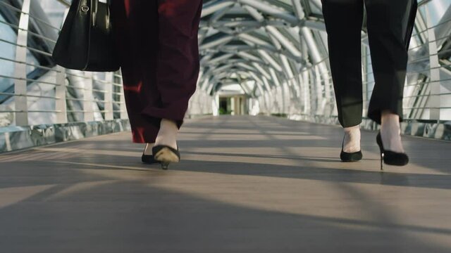 Low-section front-view slowmo of unrecognizable heels of businesswomen walking along indoor glass walkway of modern high-class office building