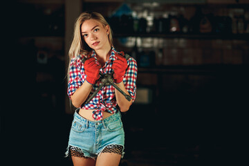 A girl in a bright plaid shirt and short denim shorts holds a balloon wrench at the service station. Handsome mechanic at work. Portrait of a beautiful model in a car garage.