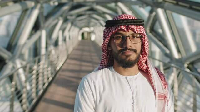 Slowmo waist-up portrait of young wealthy sheikh in traditional clothing looking at camera standing at indoor glass walkway of modern high-class office building