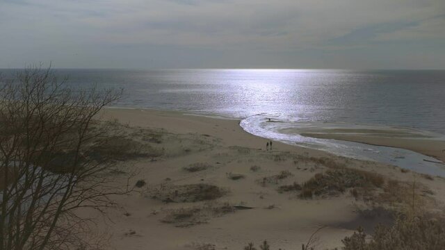 The river flows into the Baltic Sea at Saulkrasti. People walk along the yellow sand of the beach on a sunny warm spring day. Trees grow on a tall white dune in Latvia.