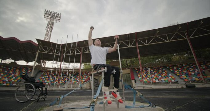 Paralympic disabled man with non-functioning legs trains sitting on a seat mount throwing a weight at a distance in the stadium