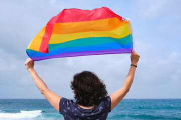 Woman holding and raising a rainbow flag over the blue sky 