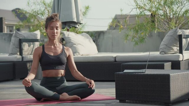 Long shot of young Caucasian woman wearing sport clothing, sitting in lotus position with eyes closed on yoga mat on roof terrace, stretching neck, then using laptop computer