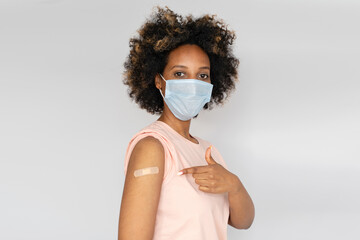 Young african american woman in medical mask showing her arm with band aid after receiving first...