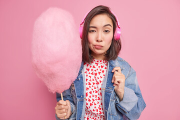 Serious attractive Asian woman looks confidently at camera holds candy floss listens music via headphones dressed in stylish clothes isolated over pink background. Teenage lifestyle concept.