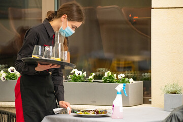 Pretty waitress with a mask clearing up, cleaning and disinfecting the table of an outdoor bar, cafe or restaurant, reopening after lockdown and restrictions © Michele Ursi
