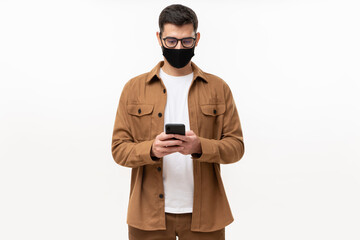 Young man in casual brown shirt and black mask, looking at his smartphone, isolated on gray background. Immunity passport online app concept