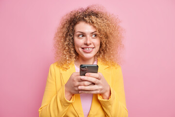 Pleased thoughtful woman with curly hair dressed in corporate clothes holds mobile phone in hands checks message box reads post online chats on social media app poses against pink background