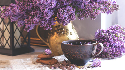 Decorative composition with a Bouquet of lilacs. Clay cup of tea and chocolate cake. A bouquet of lilacs in a vase. Spring floral background with lilacs in the interior. 
