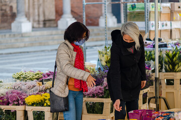 Two multiracial women with face masks in a small outdoor flower shop on a sunny day