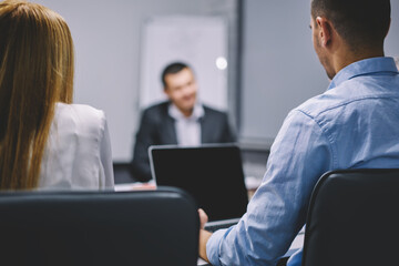 Fototapeta premium Unrecognizable male and female colleagues collaborating at office desktop with computer for online exchange, back view of professional employees brainstorming and talking about business experience