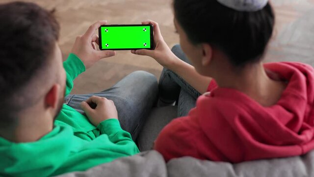 Back view of Caucasian young happy couple man and woman sitting at home and speaking online with green screen. Chroma key. Couple watching green screen in smartphone. Shot behind models shoulders.