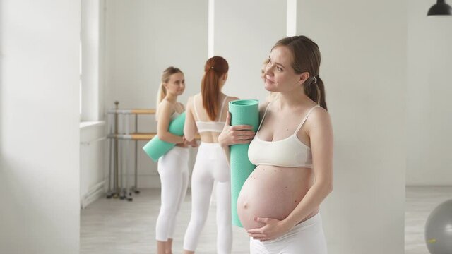 Happy mom-to-be woman with fitness mat in studio before yoga, posing, standing with naked tummy, expecting baby, preparing for giving childbirth. wellbeing concept