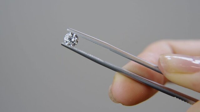 Hand with tweezers hold classic round brilliant cut diamond with grey background. Lab grown or natural diamond stone is shown by female hand holding it in the air on display in jewelry store or studio