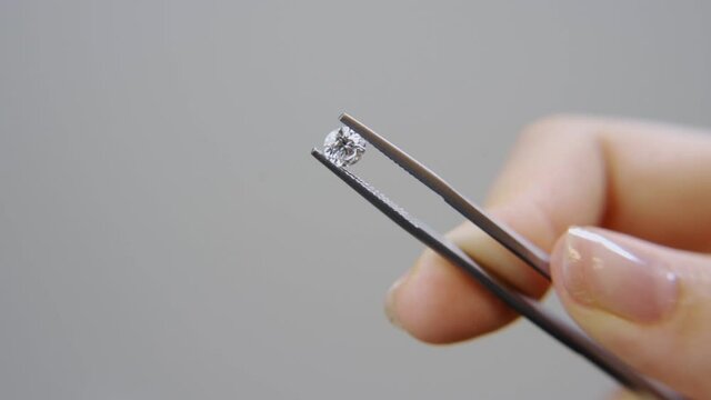 Classic round brilliant cut diamond is held with tweezers by female hand. With grey background hand is moving to show shiny stone held by specialist or professional gemologist or jeweller