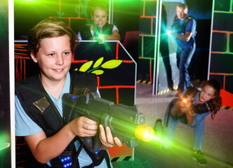 Excited cheerful positive smiling boy aiming laser gun at other players during laser tag game in dark room