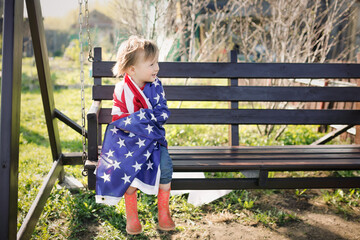 American girl Toddler child wrapped herself in the USA flag on Independence Day. A child runs with the USA flag at a picnic on July 4 on a garden swing in the yard