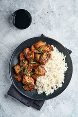 Spicy teriyaki chicken fillet pieces with rice, green onions and black sesame seeds on black plate on a grey slate, stone or concrete background. Top view with copy space.