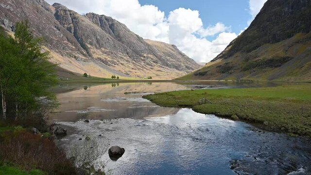 Short clip from static position of River Coe flowing with mountains either side including the Aonach Eagach ridge to the left. Accompanying tranquil audio sounds. Sunny day with blue sky and clouds.