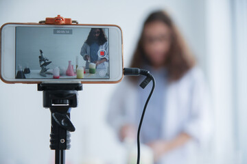 Selective focus on mobile phone on tripods is broadcasting Caucasian little girl scientist's...