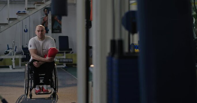 A man looks sadly intently at the camera holding boxing gloves Paralympian with defective non-functioning legs sitting in a wheelchair in an empty gym