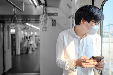 Naklejka na ściany i meble Asian man wearing face mask using smartphone on skytrain or urban train. Coronavirus (COVID-19) infection prevention in public transportation. Social distancing for pandemic protection