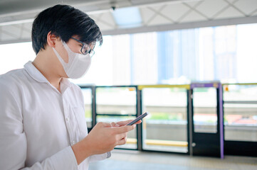 Asian man wearing face mask using smartphone at skytrain station platform. Coronavirus (COVID-19) infection prevention in public transportation. Health awareness for pandemic protection