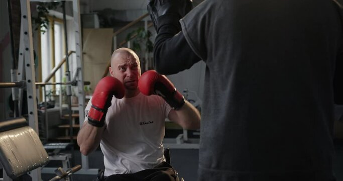 Exhausting workout in the gym Paralympian man with defective legs sitting in a wheelchair Boxing with a coach in boxing gloves
