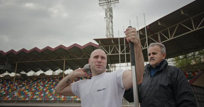 Training of a Paralympian man with non-functioning legs and his trainer preparation for throwing a weight lift for a distance on the stadium