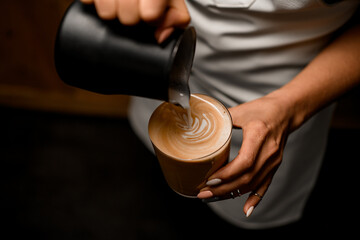 close-up of woman barista carefully drawing pattern pouring milk into glass with latte