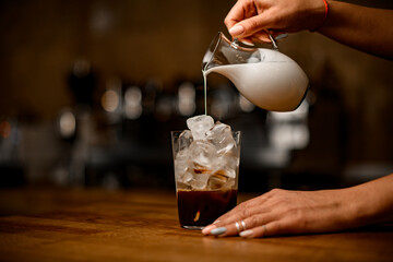 barista's hand holds jug of milk and pours it into glass with coffee and ice