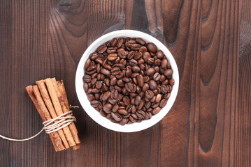 Grains of coffee in white bowl and bunch of cinnamon sticks on wooden background