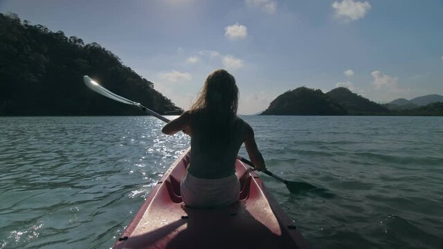 Silhouette of woman tourist raising hands while sailing in canoe along sea bay water to distant hilly island backside view. Traveling to tropical countries. Sports girl is sailing on kayak in ocean.