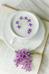 Mug of cappuccino on a beige background. Lilac flowers, book. Rest at home. Quarantine.