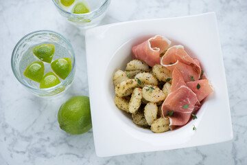 Potato gnocchi with green basil pesto and prosciutto served in a white plate, elevated view on a light-grey marble background