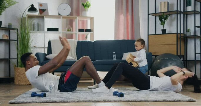Home workout concept where attractive positive active diverse couple doing press exercises with clapps on the floor while their dark-skinned kid playing near