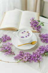 Mug of cappuccino on a beige background. Lilac flowers, book. Rest at home. Quarantine.