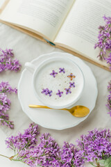 Mug of cappuccino on a beige background. Lilac flowers, book. Rest at home. Quarantine.